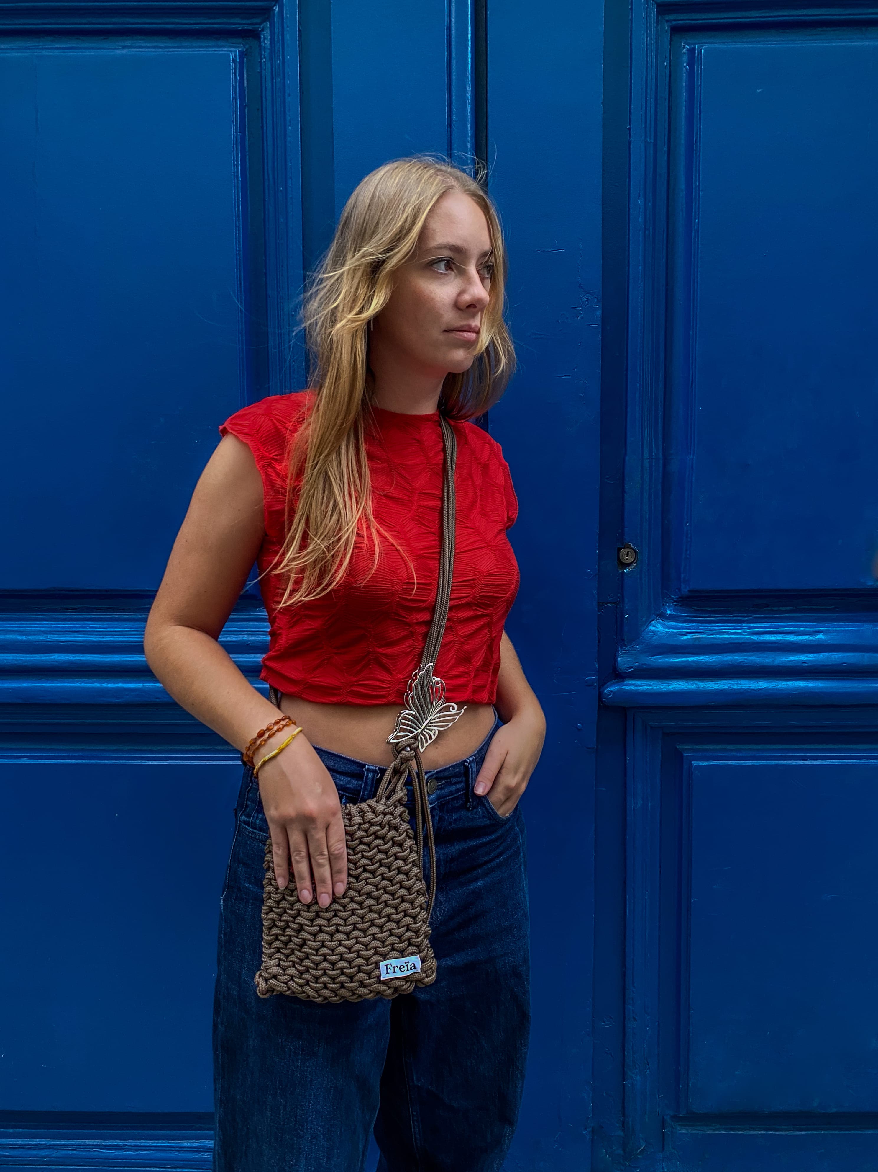 Femme devant une porte bleue avec un sac Freïa en paracorde marron et un top rouge