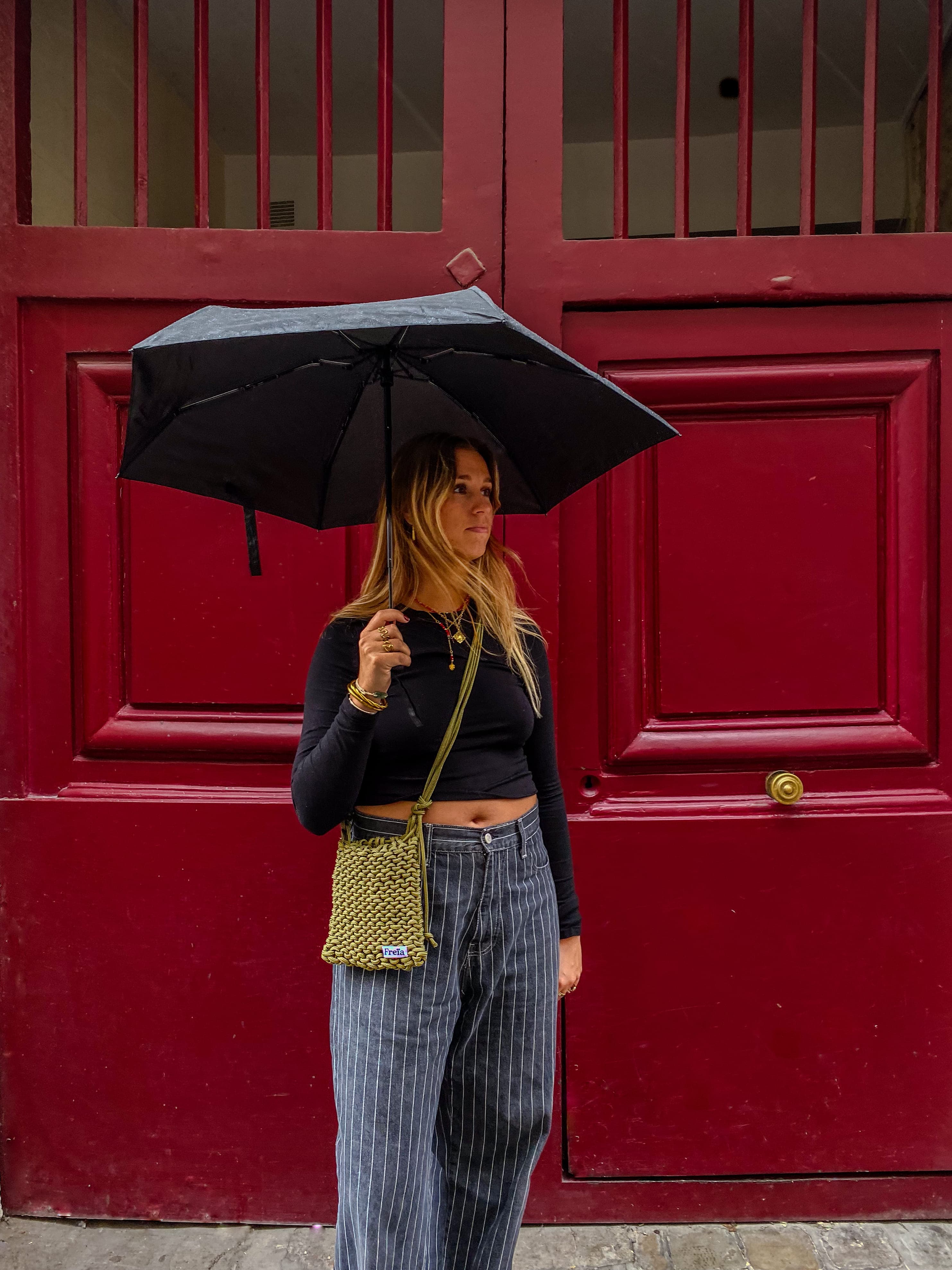 Femme tenant un parapluie devant une porte rouge avec un sac Freïa en paracorde kaki